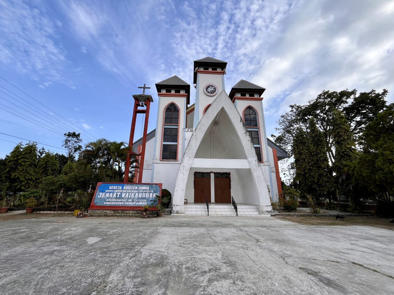 A white church with red trim and three spires stands under a blue sky, surrounded by lush green trees and a sign in front.