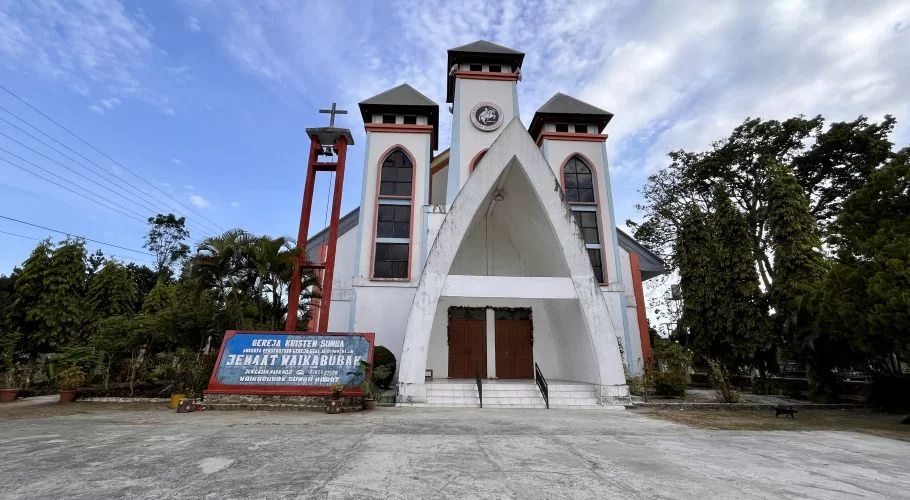 A white church with red trim and three spires stands under a blue sky, surrounded by lush green trees and a sign in front.