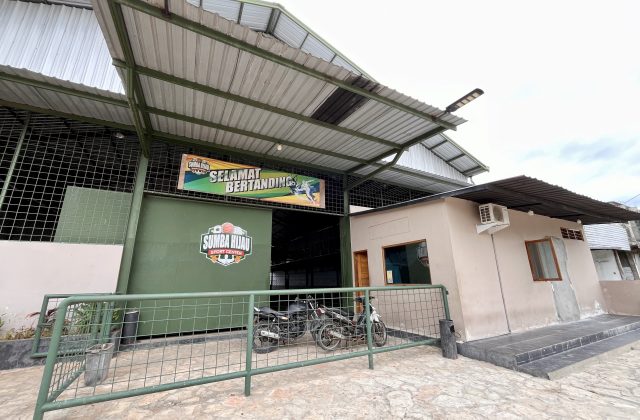 Sumba Hijau Futsal building's green entrance, displaying a "Selamat Bertanding" banner, with two motorcycles parked behind a metal fence.