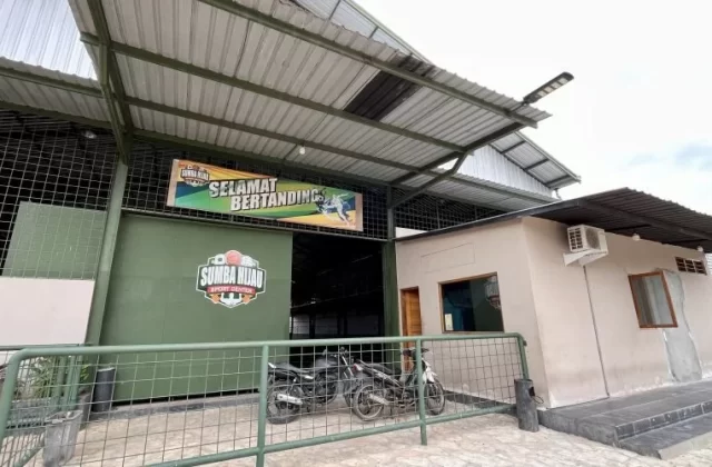 Sumba Hijau Futsal building's green entrance, displaying a "Selamat Bertanding" banner, with two motorcycles parked behind a metal fence.