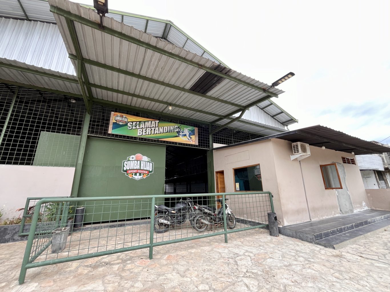 Sumba Hijau Futsal building's green entrance, displaying a "Selamat Bertanding" banner, with two motorcycles parked behind a metal fence.
