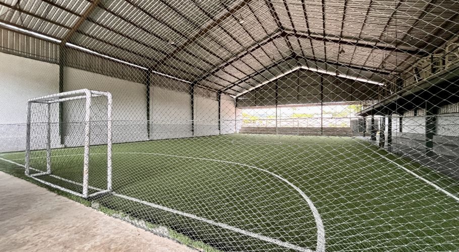 An indoor futsal court in Tambolaka, Sumba, features green artificial turf with white lines, a white goal, and a corrugated metal roof, enclosed by safety netting.