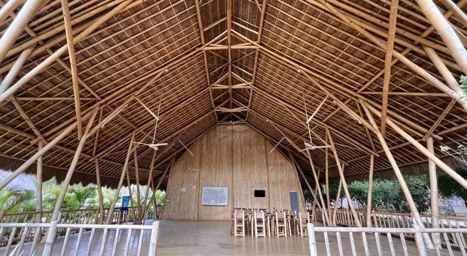 An open-air building interior features a high, peaked thatched roof and light brown bamboo structure, with a classroom setup and green foliage visible outside.