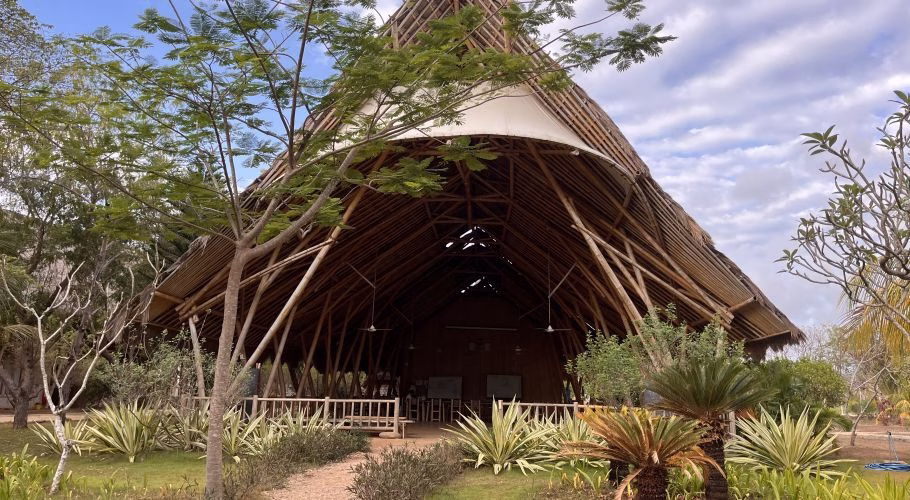 A large, open-sided building with a high bamboo and thatch roof stands amidst green tropical plants under a partly cloudy sky.