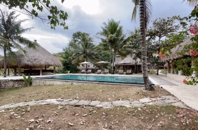 A blue outdoor swimming pool with sun loungers is surrounded by thatched-roof bungalows and green palm trees on a hillside.