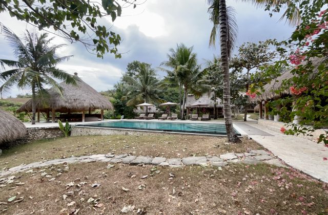 A blue outdoor swimming pool with sun loungers is surrounded by thatched-roof bungalows and green palm trees on a hillside.