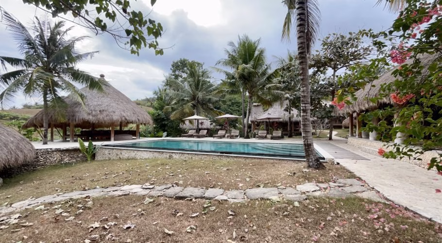 A blue outdoor swimming pool with sun loungers is surrounded by thatched-roof bungalows and green palm trees on a hillside.