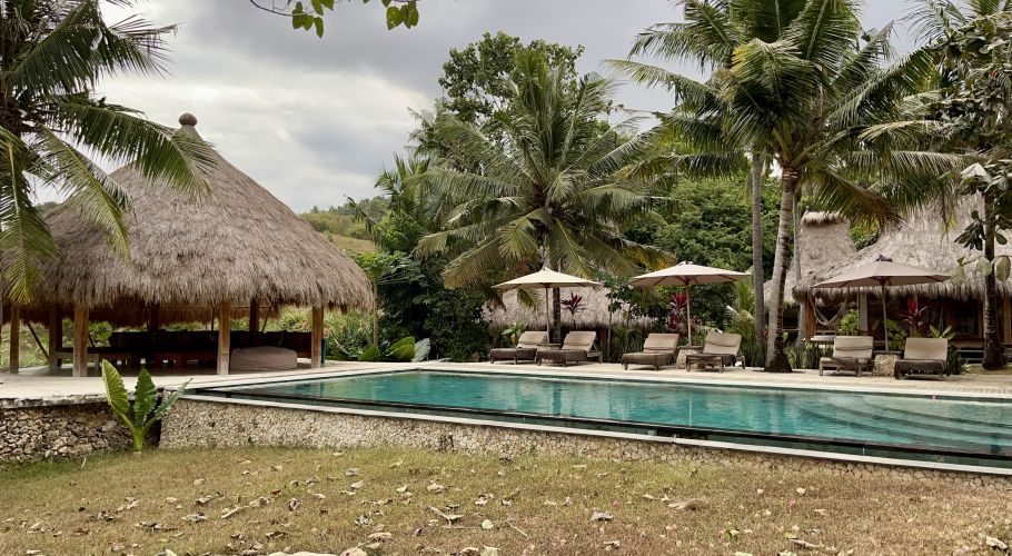 A tranquil outdoor swimming pool is bordered by sun loungers, thatched-roof bungalows, and lush palm trees under an overcast sky.