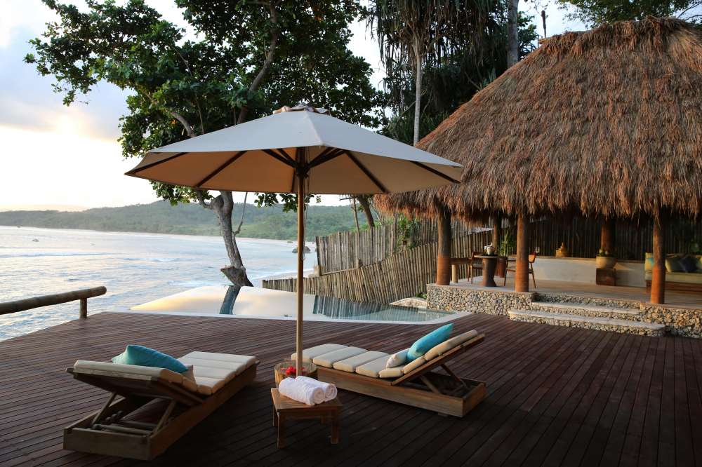 A wooden deck with two lounge chairs, an umbrella, and an infinity pool overlooks a calm ocean and distant green hills at sunset.
