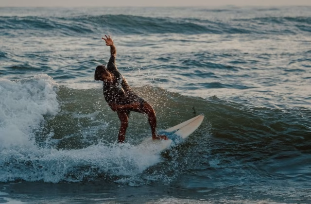 A surfer in a dark wetsuit rides a breaking wave on a white board, with water splashing under a hazy sky.