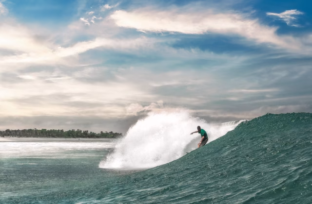 A surfer in a green shirt rides a powerful, breaking blue-green wave, with white spray and a lush tropical coastline under a cloudy sky.