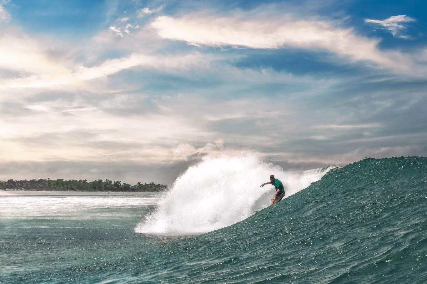 A surfer in a green shirt rides a powerful, breaking blue-green wave, with white spray and a lush tropical coastline under a cloudy sky.