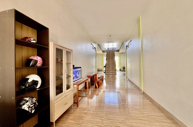 A bright, clean guest house lobby features a dark shelf with colorful helmets, an aquarium, and a long tiled hallway leading to an upstairs staircase.