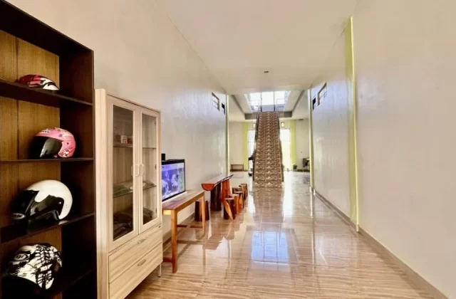 A bright, clean guest house lobby features a dark shelf with colorful helmets, an aquarium, and a long tiled hallway leading to an upstairs staircase.