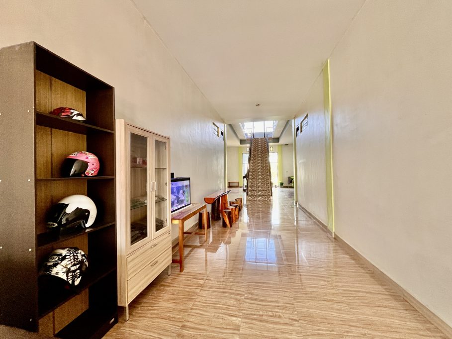 A bright, clean guest house lobby features a dark shelf with colorful helmets, an aquarium, and a long tiled hallway leading to an upstairs staircase.