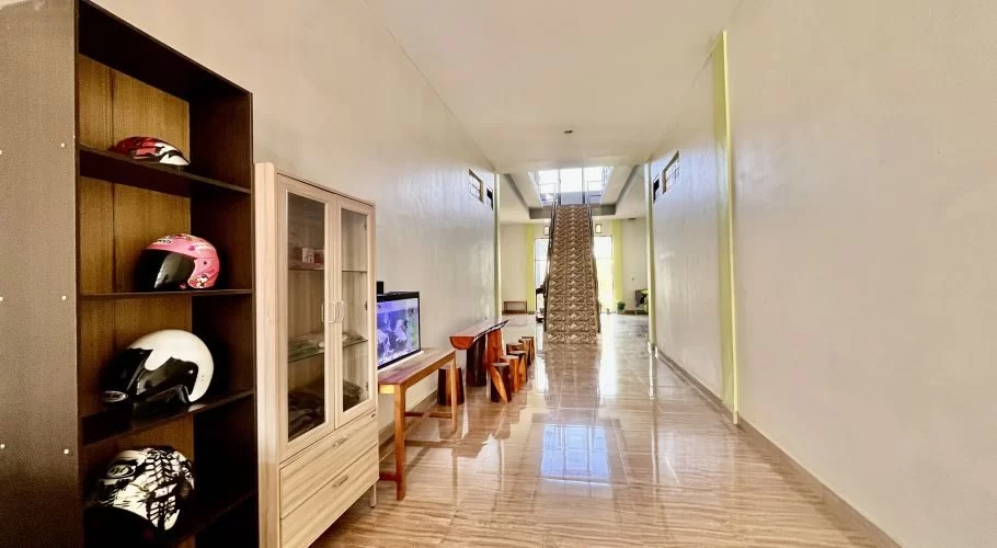 A bright, clean guest house lobby features a dark shelf with colorful helmets, an aquarium, and a long tiled hallway leading to an upstairs staircase.