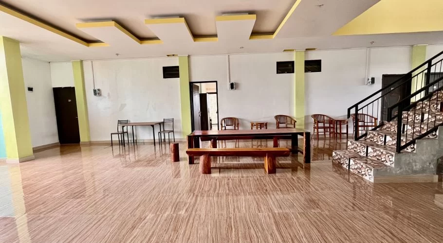 A clean, spacious guest house lobby features shiny brown tiled floors, white walls with green accents, dark wood furniture, and a patterned staircase leading upstairs.