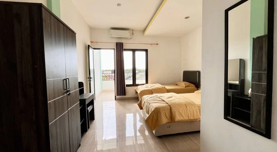 A clean, modern guest room features two single beds with light brown bedding, a dark wardrobe, a desk, and a balcony.