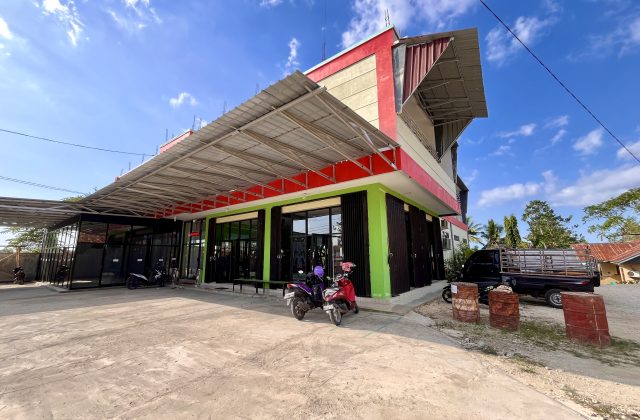 A modern two-story building, Surya Baru Gym, with red, cream, and green walls, glass windows, and a wide metal awning, under a blue sky.