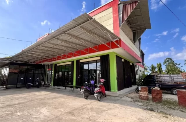 A modern two-story building, Surya Baru Gym, with red, cream, and green walls, glass windows, and a wide metal awning, under a blue sky.