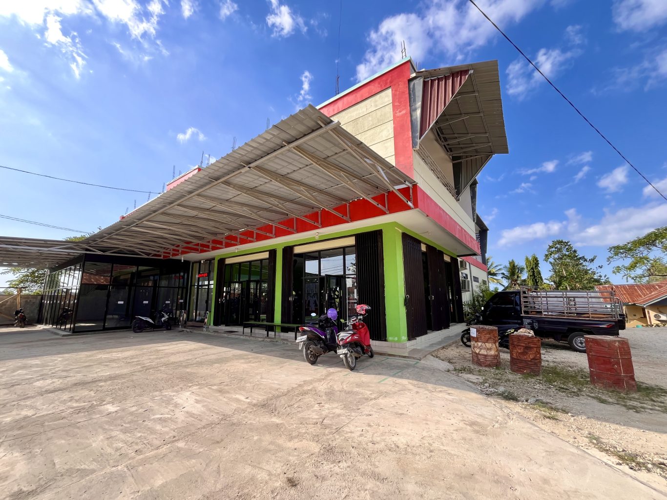 A modern two-story building, Surya Baru Gym, with red, cream, and green walls, glass windows, and a wide metal awning, under a blue sky.