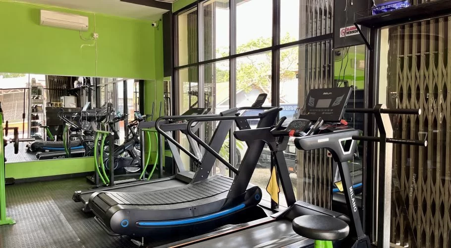 A well-lit gym features bright green walls, black rubber flooring, large mirrors, and several new treadmills and exercise bikes.
