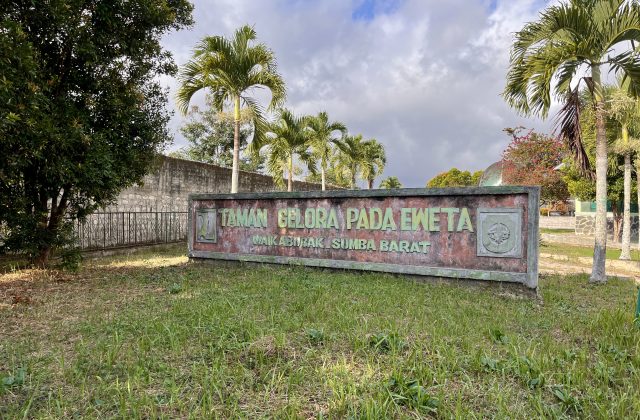 A weathered sign for Taman Gelora Pada Eweta in Waikabubak, Sumba Barat, stands amidst green grass and tall palm trees under a cloudy sky.