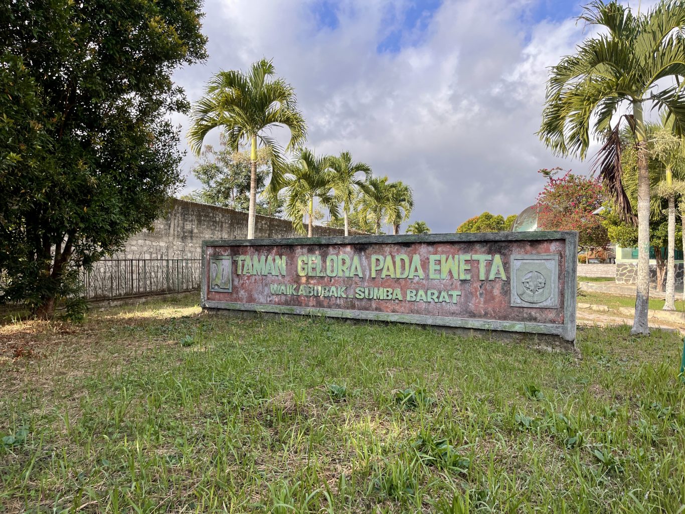 A weathered sign for Taman Gelora Pada Eweta in Waikabubak, Sumba Barat, stands amidst green grass and tall palm trees under a cloudy sky.