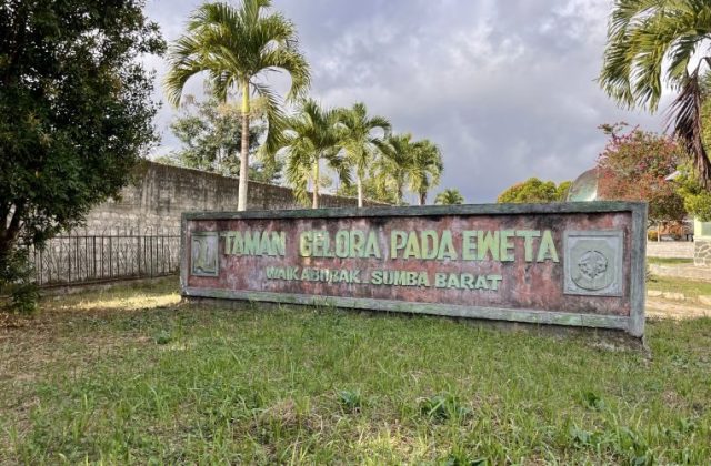A weathered sign for Taman Gelora Pada Eweta in Waikabubak, Sumba Barat, stands amidst green grass and tall palm trees under a cloudy sky.