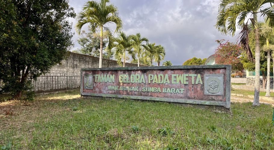 A weathered sign for Taman Gelora Pada Eweta in Waikabubak, Sumba Barat, stands amidst green grass and tall palm trees under a cloudy sky.