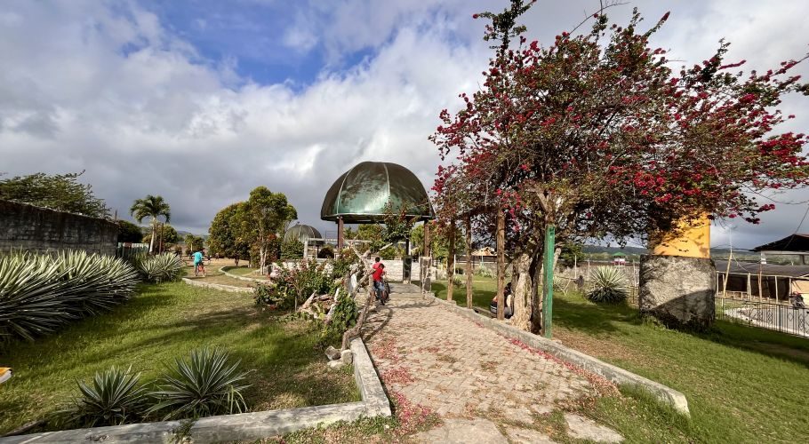 A lush green park features paved paths, a red-flowered bougainvillea tree, and two dome-shaped gazebos under a partly cloudy sky, with a person cycling in the distance.