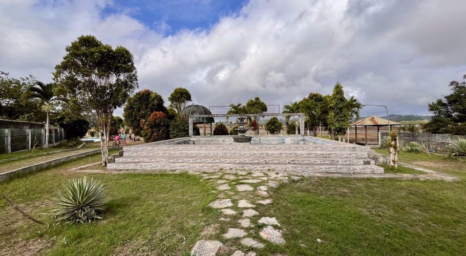 A wide view of Taman Gelora Pada Eweta park shows an empty tiered fountain surrounded by green grass, diverse trees, and a stone path under a cloudy sky, with children on bikes in the distance.