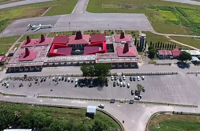 An aerial view shows Tambolaka Sumba Airport with its distinctive red-roofed terminal, a parked airplane on the tarmac, and a busy car park, surrounded by green fields.