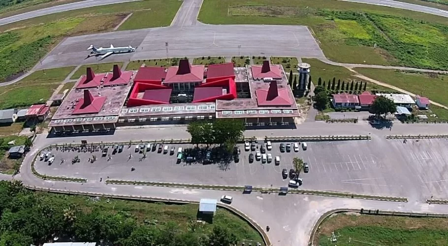 An aerial view shows Tambolaka Sumba Airport with its distinctive red-roofed terminal, a parked airplane on the tarmac, and a busy car park, surrounded by green fields.