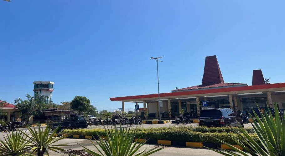 Tambolaka Sumba Airport terminal with a red roof, two tall red structures, a control tower, and a parking lot under a clear blue sky.