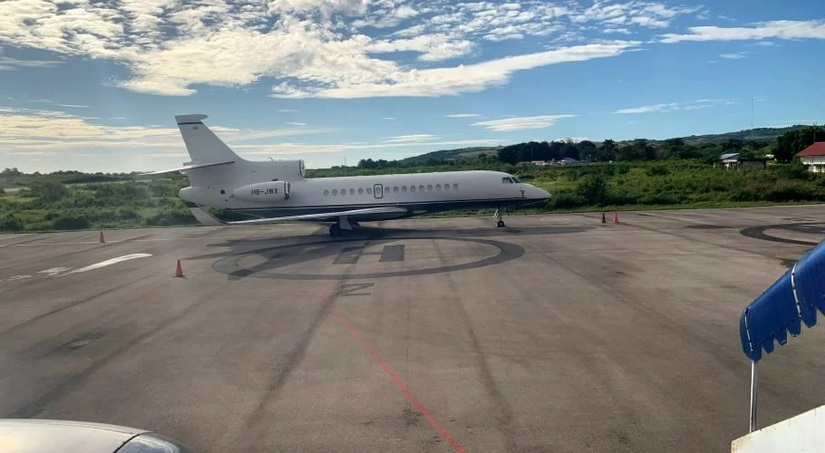 A white private jet is parked on an asphalt tarmac at Tambolaka Sumba Airport, surrounded by green vegetation under a partly cloudy blue sky.