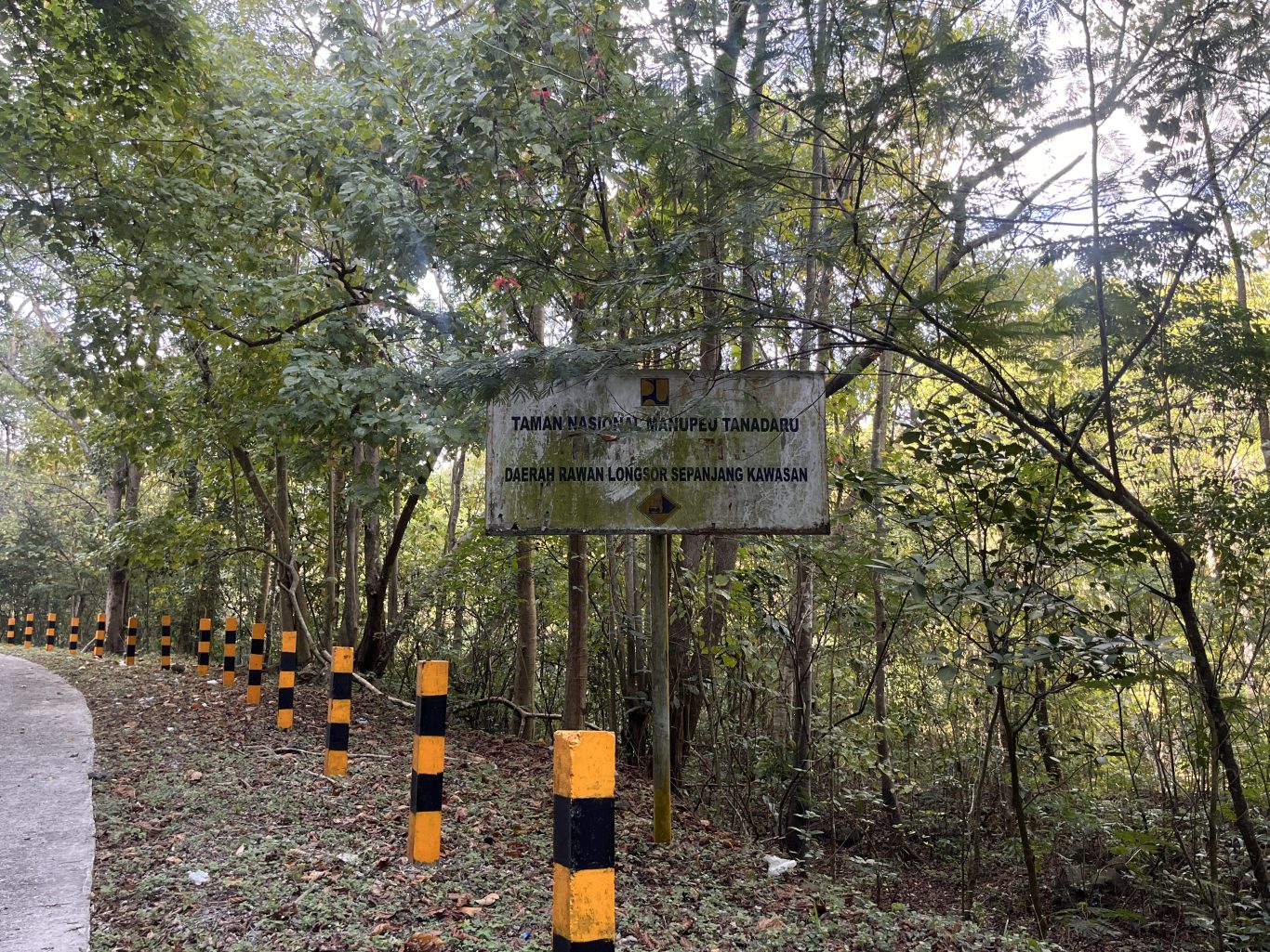 A weathered sign for Manupeu Tanadaru National Park stands amidst dense green forest, with a paved road and yellow and black striped posts on the left.