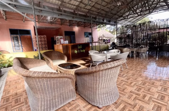 An outdoor cafe seating area features wicker chairs and tables on a tiled floor, with a peach building and a wooden reception desk visible under a metal roof.