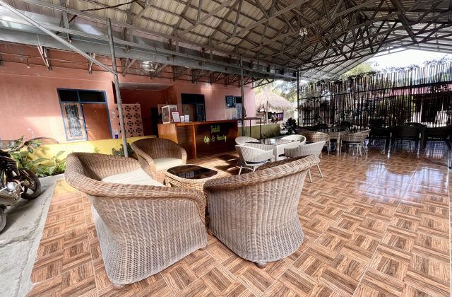 An outdoor cafe seating area features wicker chairs and tables on a tiled floor, with a peach building and a wooden reception desk visible under a metal roof.