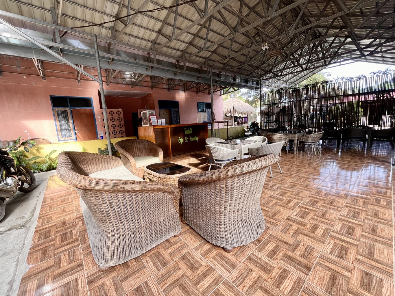 An outdoor cafe seating area features wicker chairs and tables on a tiled floor, with a peach building and a wooden reception desk visible under a metal roof.
