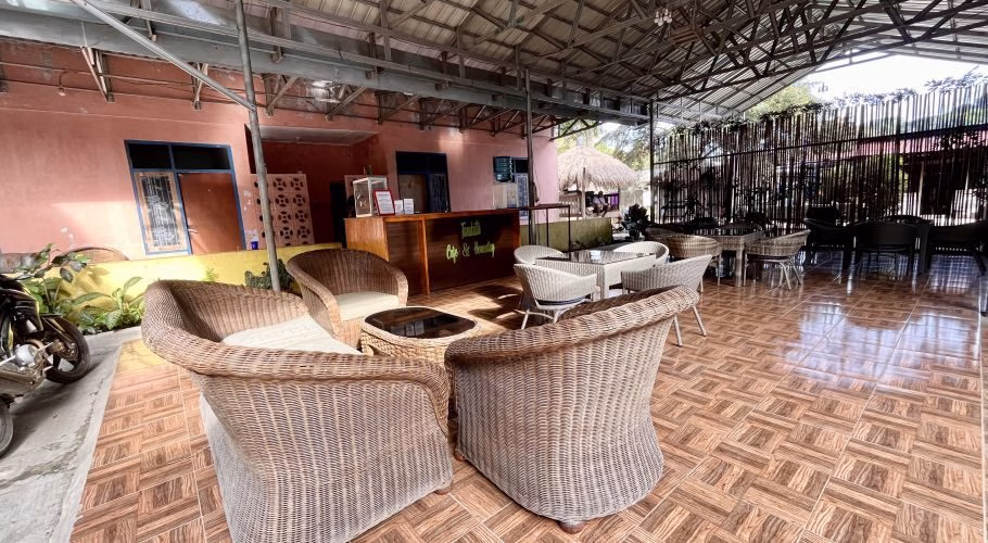 An outdoor cafe seating area features wicker chairs and tables on a tiled floor, with a peach building and a wooden reception desk visible under a metal roof.