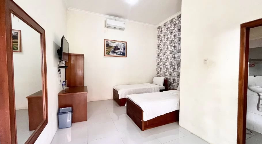 A clean, simple homestay room features two single beds with white linens, a wooden dresser with a TV, and a mirror, all against white walls and a patterned accent wall.