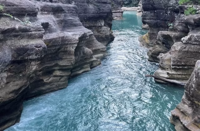 A narrow river with vibrant turquoise water flows between tall, layered grey rock formations in a natural canyon.