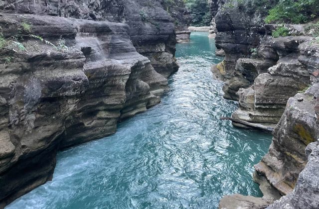 A narrow river with vibrant turquoise water flows between tall, layered grey rock formations in a natural canyon.