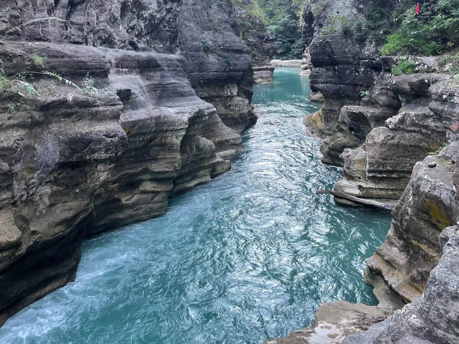 A narrow river with vibrant turquoise water flows between tall, layered grey rock formations in a natural canyon.