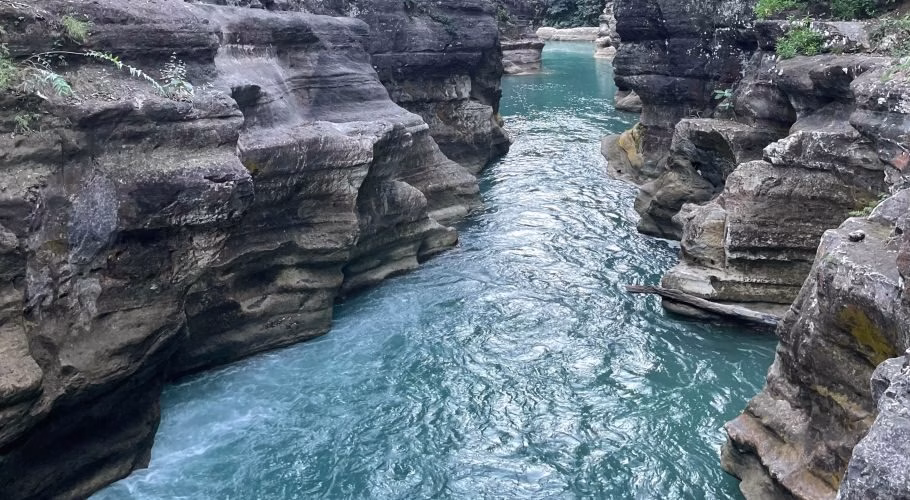 A narrow river with vibrant turquoise water flows between tall, layered grey rock formations in a natural canyon.