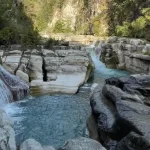 Tanggedu Waterfall cascades over light-colored rock formations into multiple turquoise pools within a rocky gorge surrounded by green foliage.