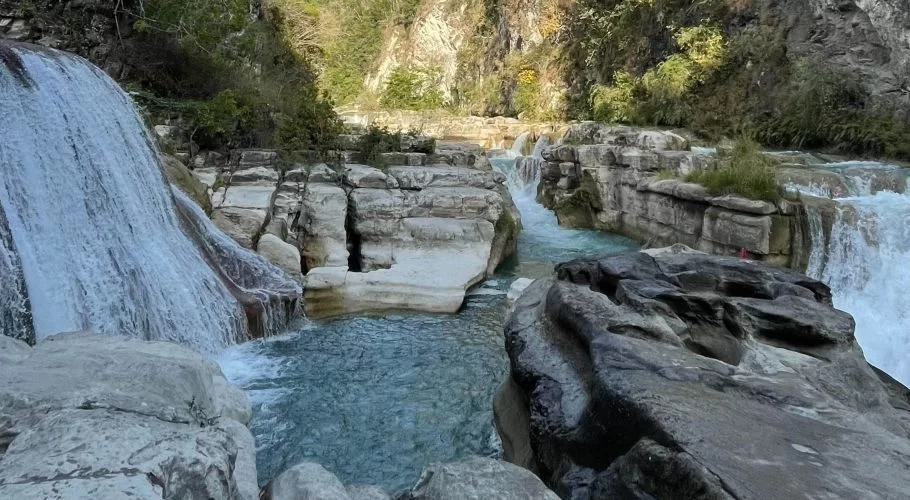 Tanggedu Waterfall cascades over light-colored rock formations into multiple turquoise pools within a rocky gorge surrounded by green foliage.