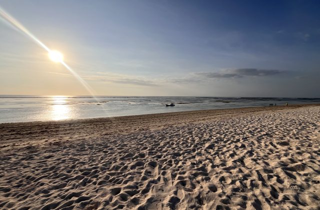 A wide, pristine white sand beach stretches towards calm, reflective turquoise-green waters under a bright sun with a lens flare. A small boat floats in the distance, and faint figures walk along the shore.