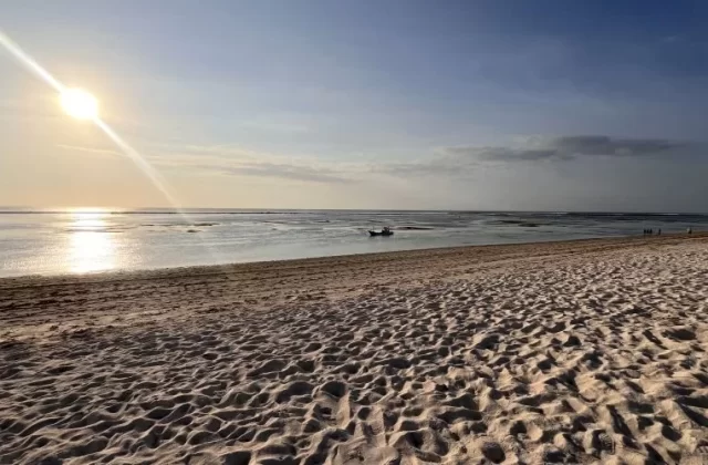 A wide, pristine white sand beach stretches towards calm, reflective turquoise-green waters under a bright sun with a lens flare. A small boat floats in the distance, and faint figures walk along the shore.